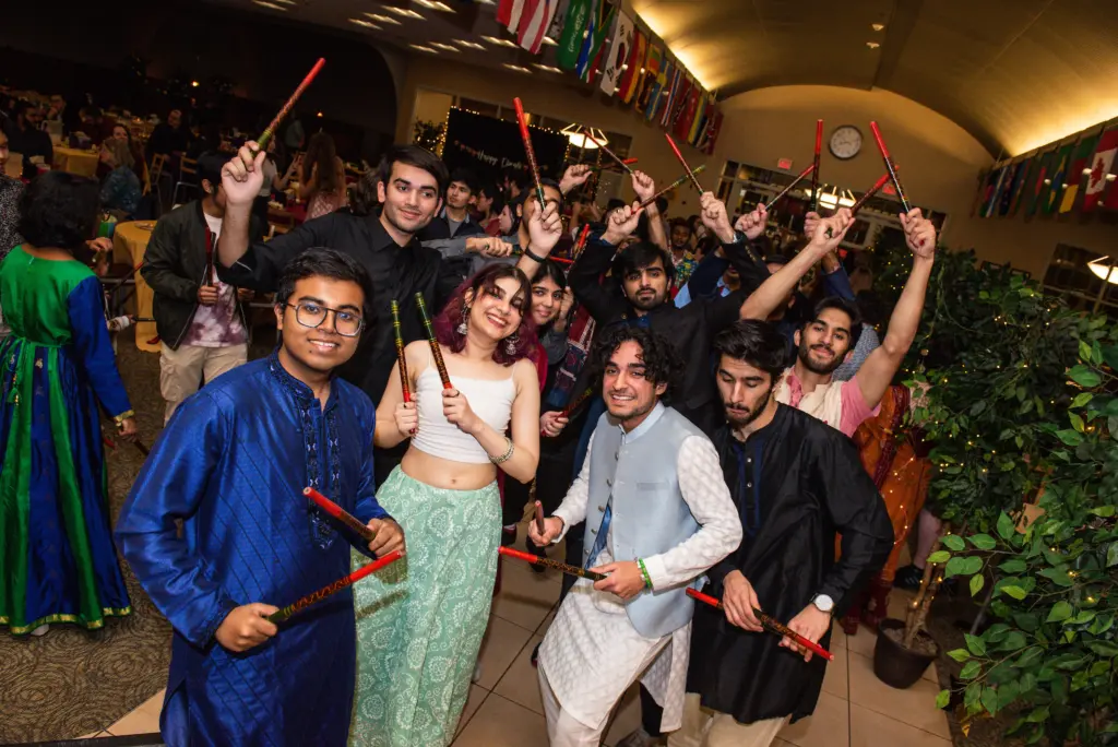 A group of young people in colorful traditional South Asian attire smile and pose with dandiya sticks raised in a festive indoor setting decorated with international flags.