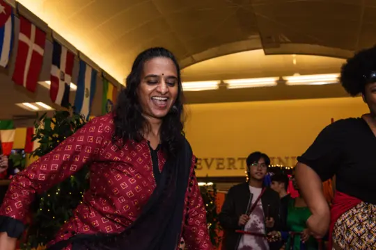 A woman in a red patterned dress smiles and dances at an indoor event decorated with international flags, while others watch or participate in the background.