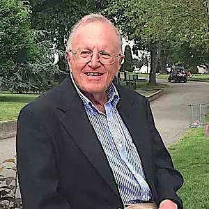 An older man with glasses and a friendly smile sits outdoors on a bench, wearing a dark jacket over a light blue shirt. Trees and greenery are visible in the background along a park path.
