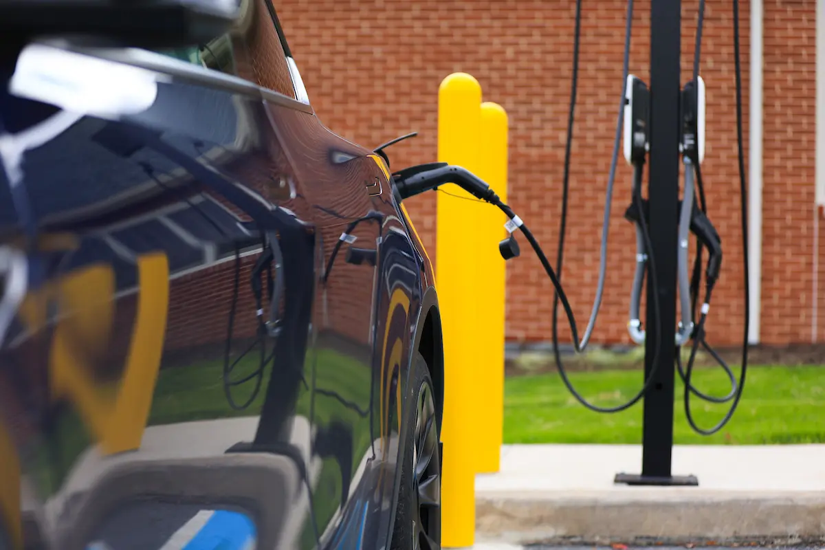 A close-up of an electric vehicle plugged into a charging station outdoors, with bright yellow bollards and a brick wall in the background.