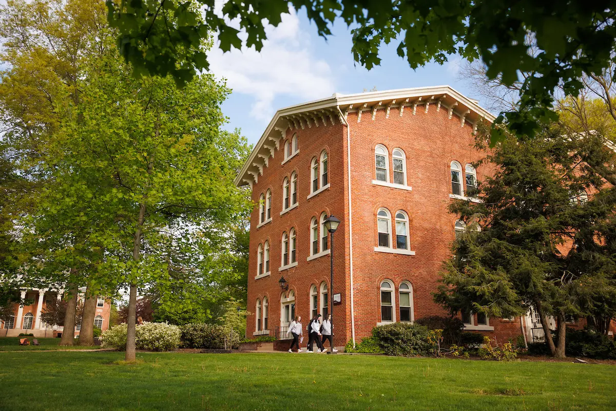 A historic red brick building with white trim sits among green trees and a manicured lawn, as three people—perhaps members of Our Leadership—walk on a path in front of it under a partly cloudy sky.