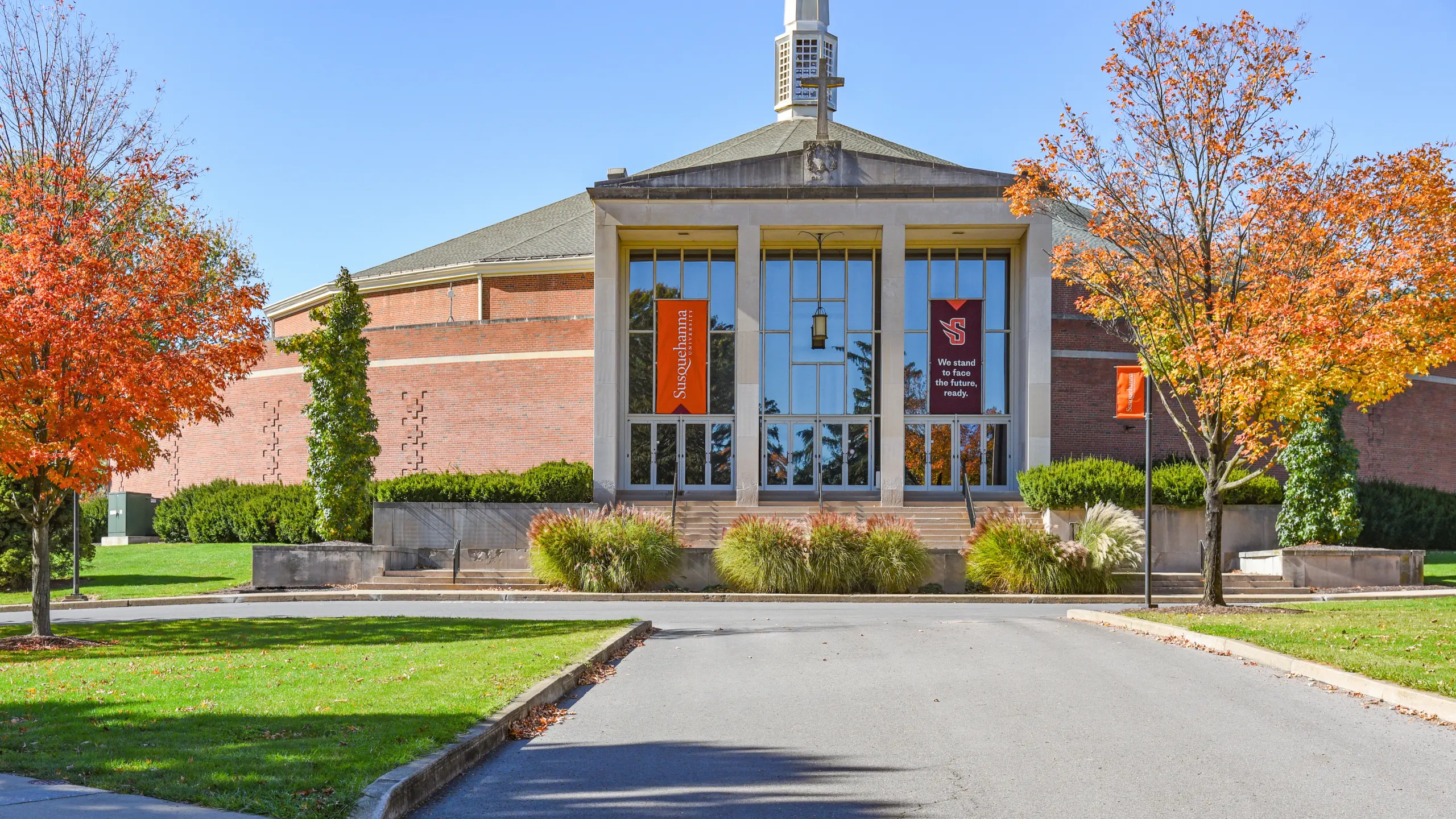 A modern brick building with large glass windows, surrounded by autumn trees with orange leaves; steps and landscaped bushes lead up to the entrance, offering a welcoming glimpse into vibrant campus life.