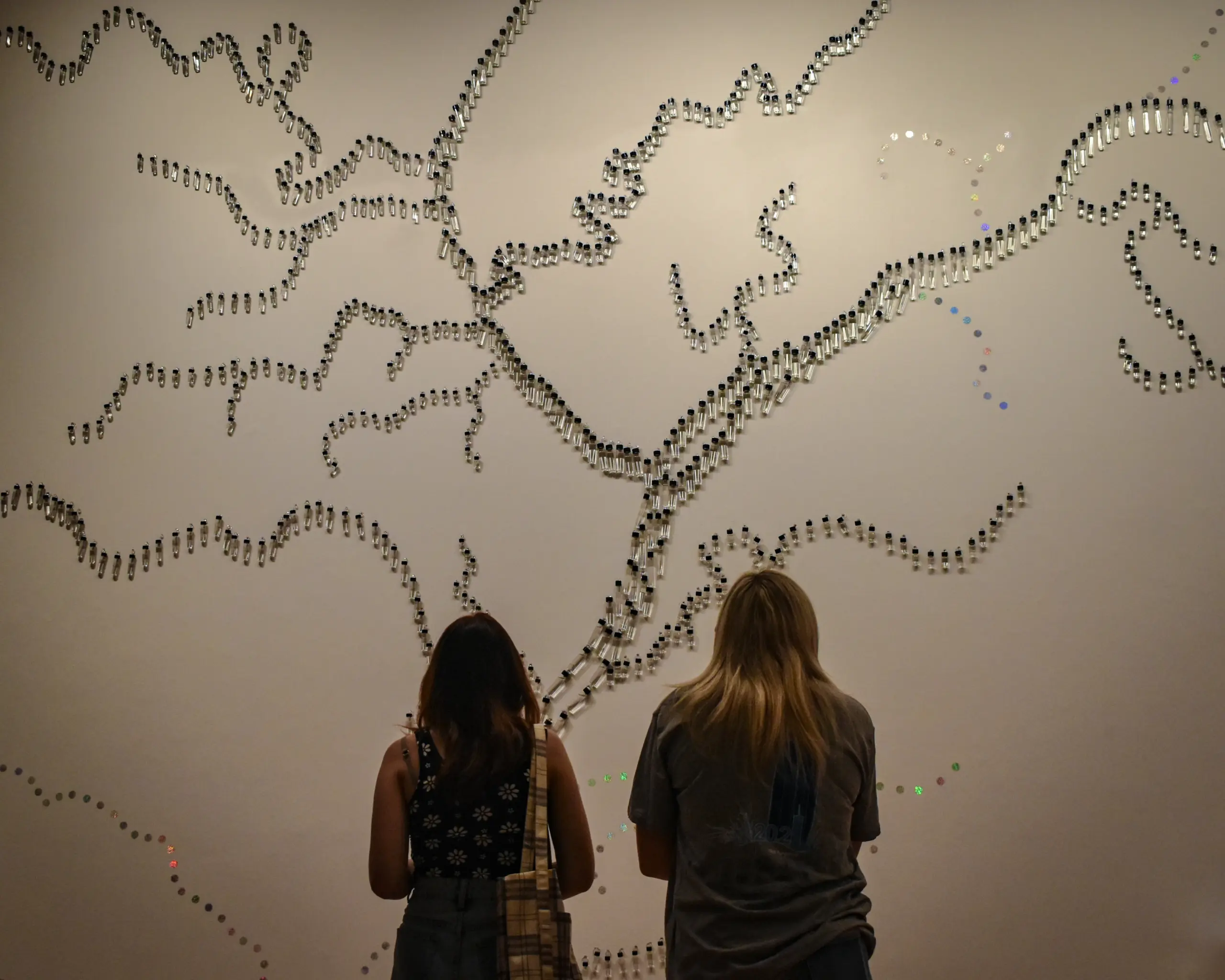 Two people stand facing a wall with an abstract installation made of small objects arranged in winding, branching patterns resembling a network or map. The lighting casts soft shadows, creating a contemplative atmosphere.
