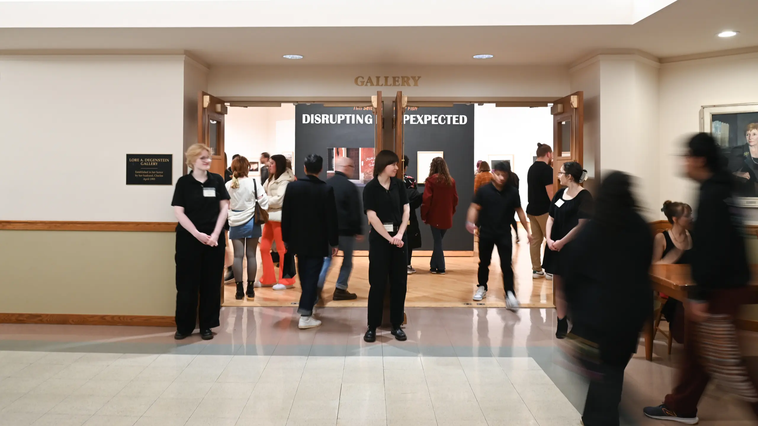 A group of people stand and walk near the entrance of an art gallery titled 