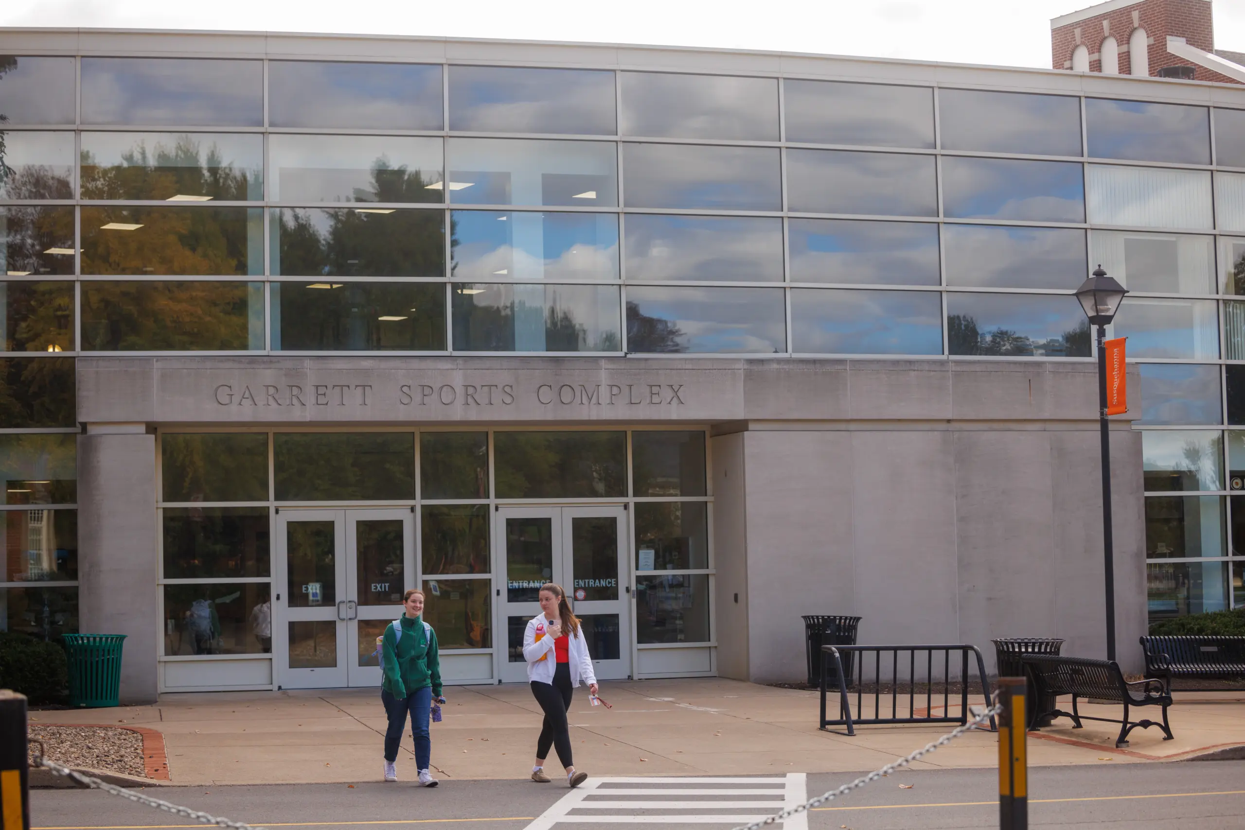 Two people walk out of the Garrett Sports Complex, a modern building with large glass windows, on a cloudy day. Trees and reflections of the sky are visible in the windows.