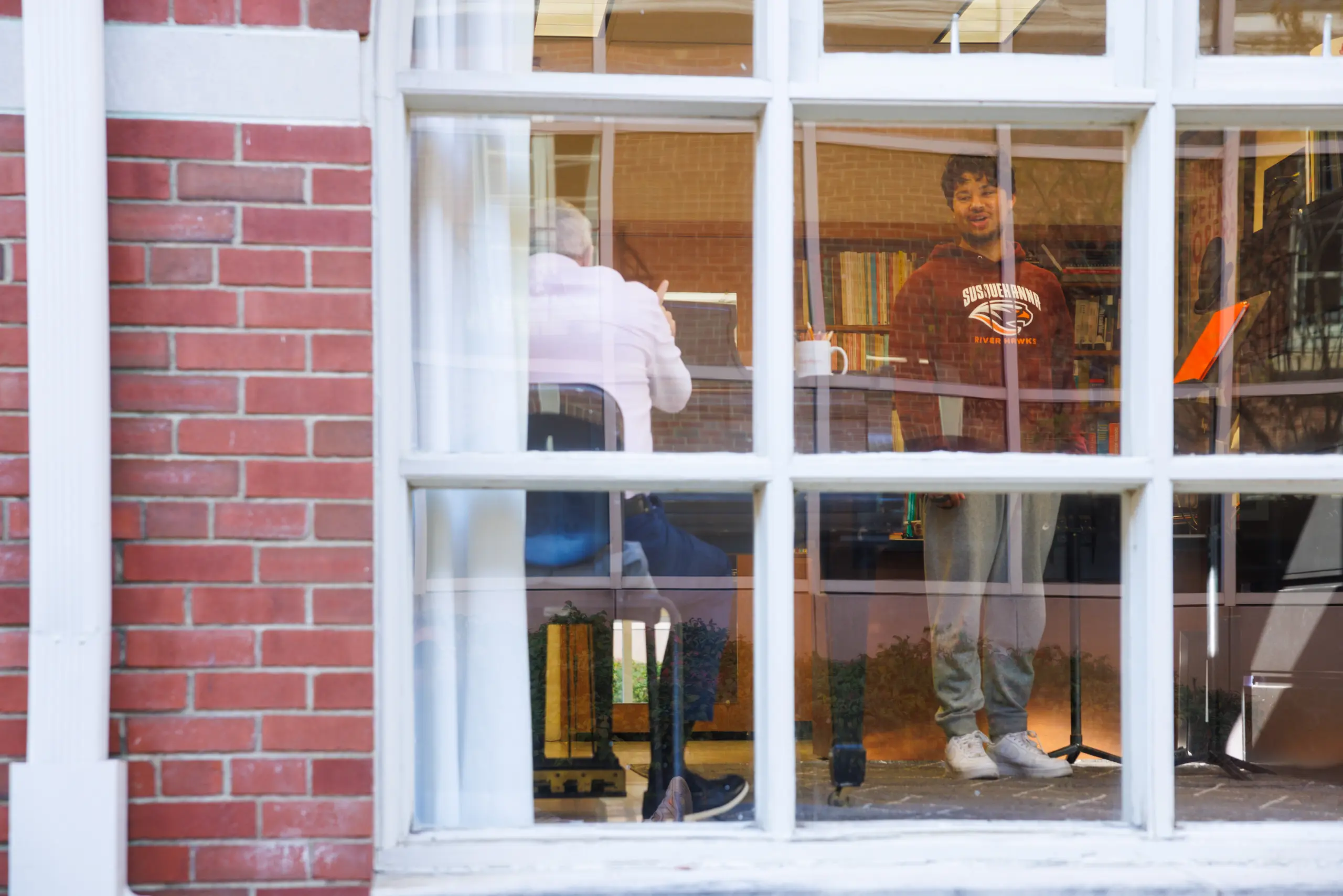 Two people are inside a room with large windows; one is sitting at a desk with their back to the window, while the other stands facing forward, wearing a sweatshirt and smiling. Bookshelves are visible in the background.