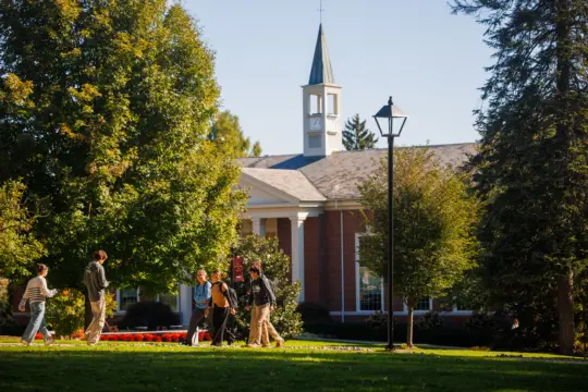 Students walk and talk on a grassy campus, surrounded by trees, with a brick building and a white steeple in the background on a sunny day.