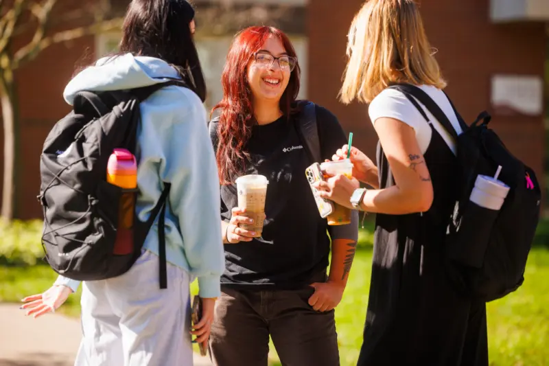 Three students with backpacks and drinks stand outside, smiling and talking together on a sunny day. One student has bright red hair and glasses, and all appear relaxed and happy.