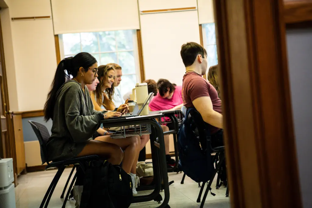Several students, including transfer students, sit at desks in a classroom, focused on their laptops and notebooks. Sunlight streams through the windows behind them, and backpacks rest on the floor by their chairs.