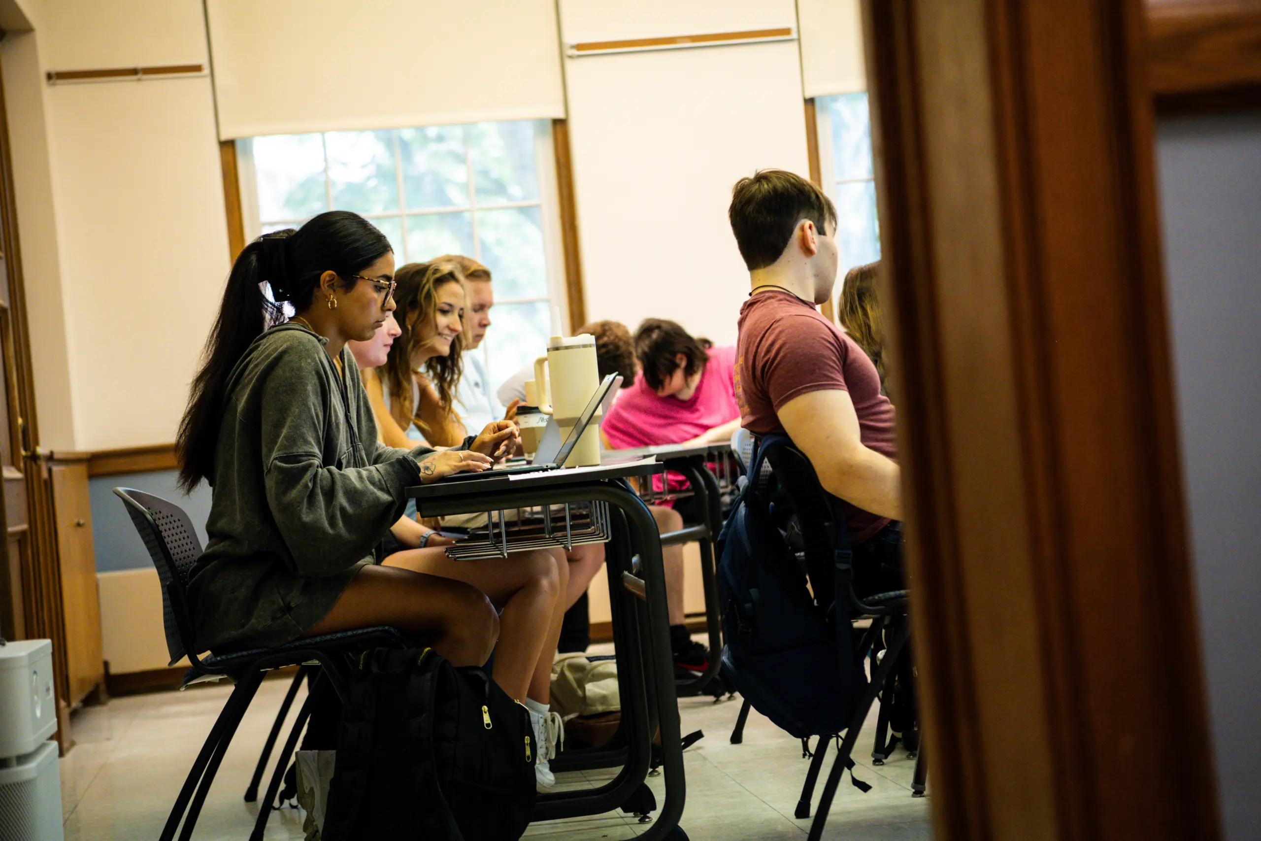 Several students, including transfer students, sit at desks in a classroom, focused on their laptops and notebooks. Sunlight streams through the windows behind them, and backpacks rest on the floor by their chairs.
