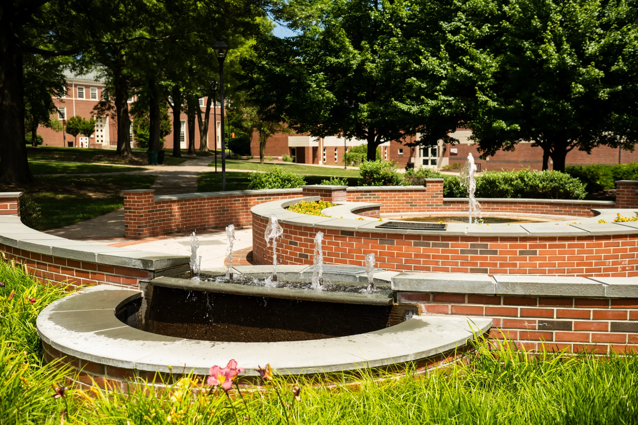 A brick fountain with multiple water jets is surrounded by greenery and trees on a sunny day, with buildings visible in the background.
