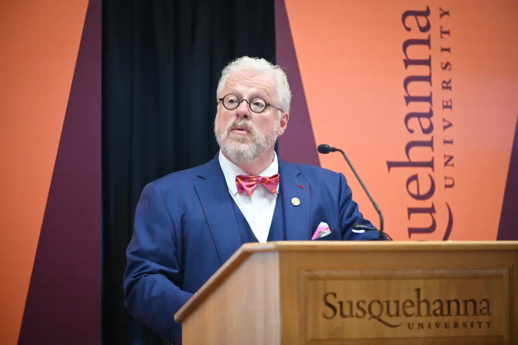 A man with white hair and a beard, representing Our Leadership, wears glasses, a blue suit, and a red bow tie as he speaks at a podium labeled 