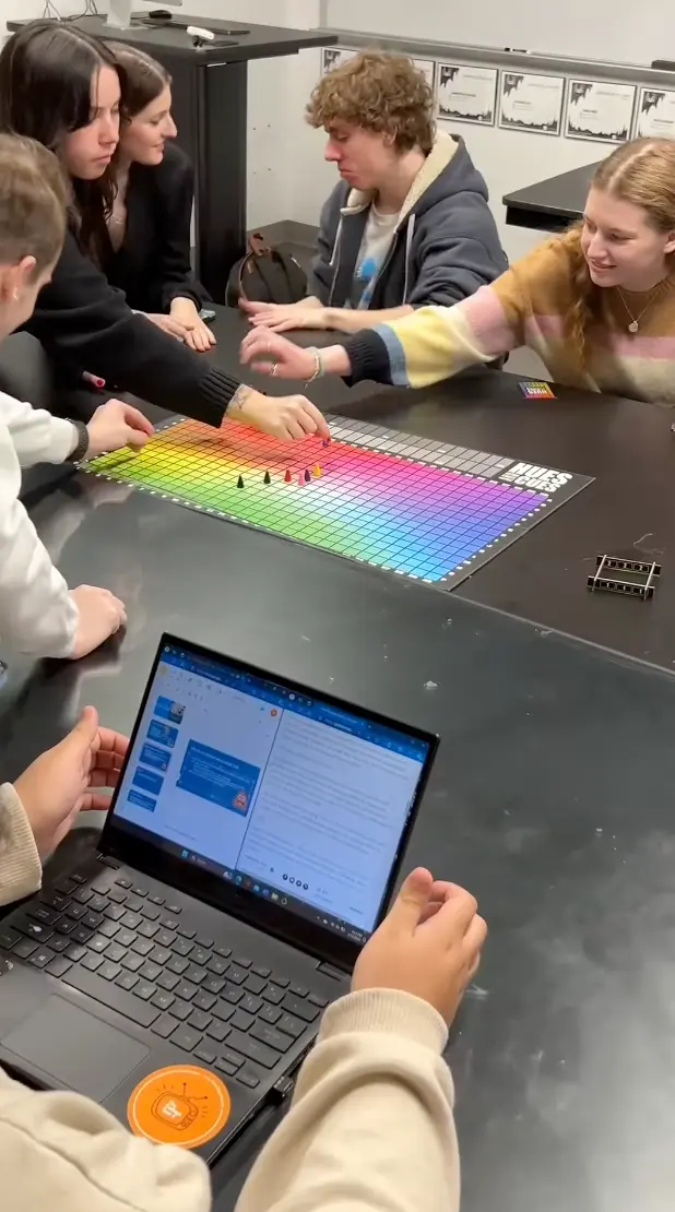 Five academics sit around a table playing a colorful board game, with one person reaching over the board. A laptop with a presentation open is in the foreground. The lively scene appears to be set in a classroom or lab.