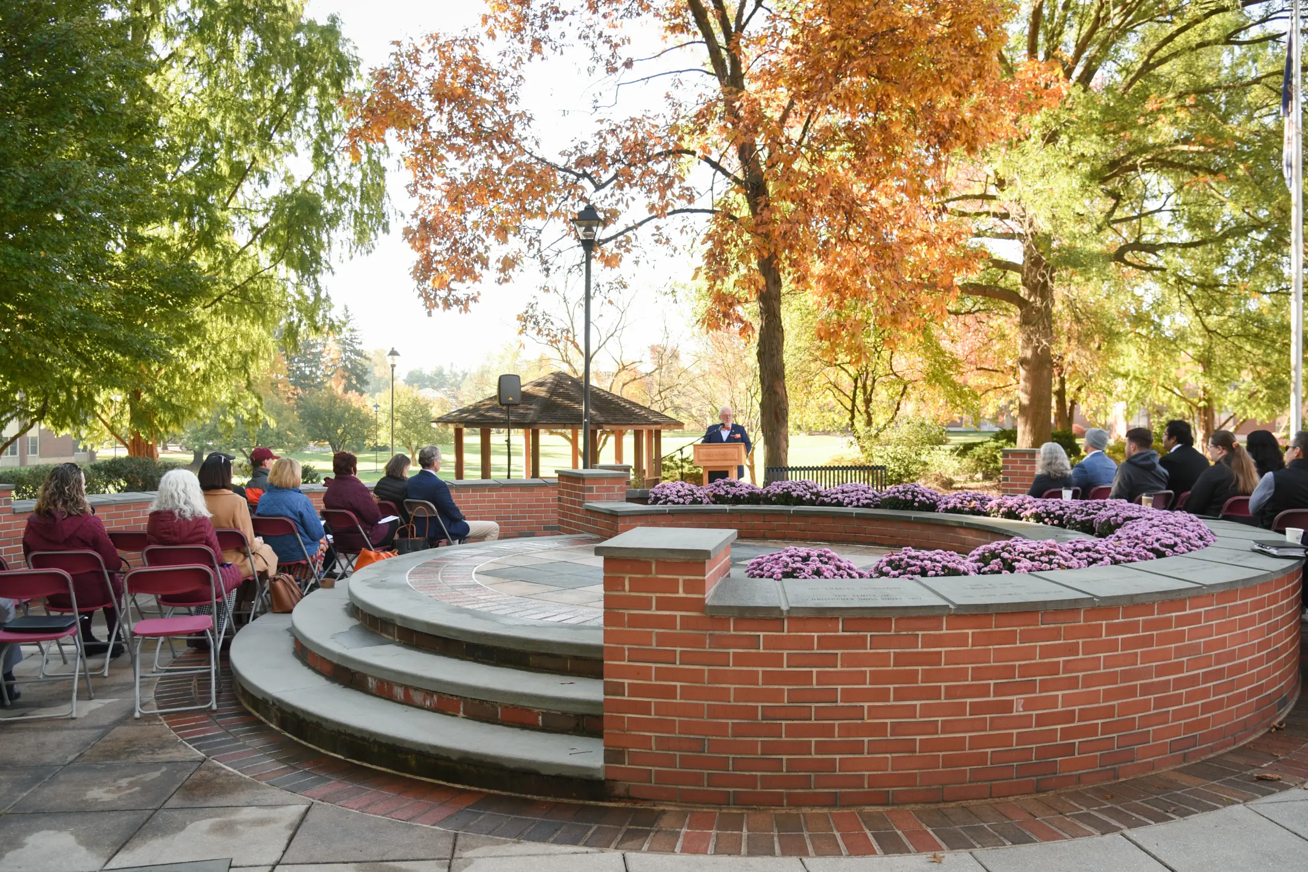 A group of people sitting on chairs outdoors attends a formal event by a circular brick structure with purple flowers. A person stands at a podium under autumn trees, with a small wooden gazebo in the background.