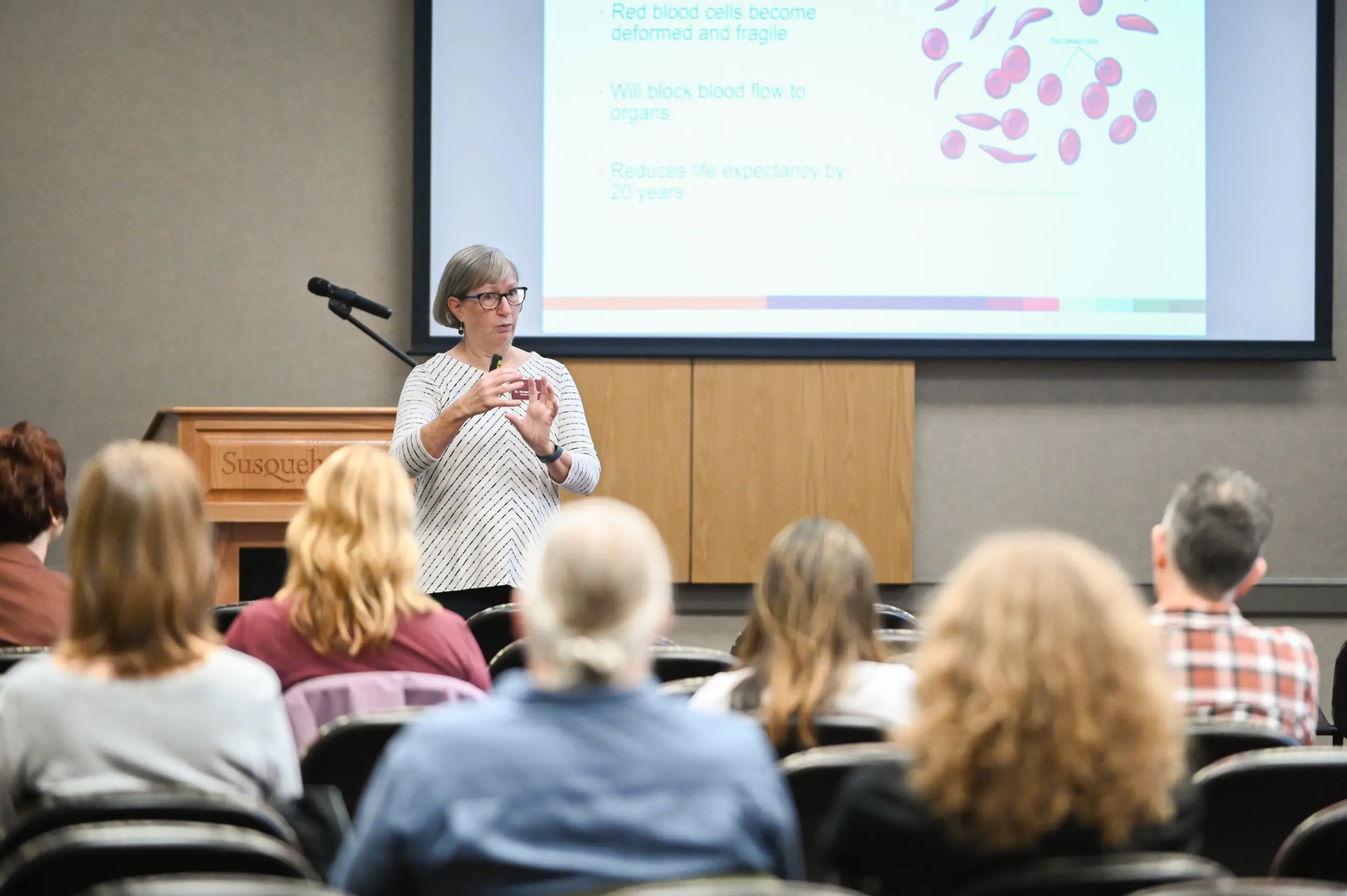 A woman gives a presentation to an audience in a classroom, standing in front of a screen displaying information about red blood cells and a diagram about their structure. The audience is seated and listening attentively.