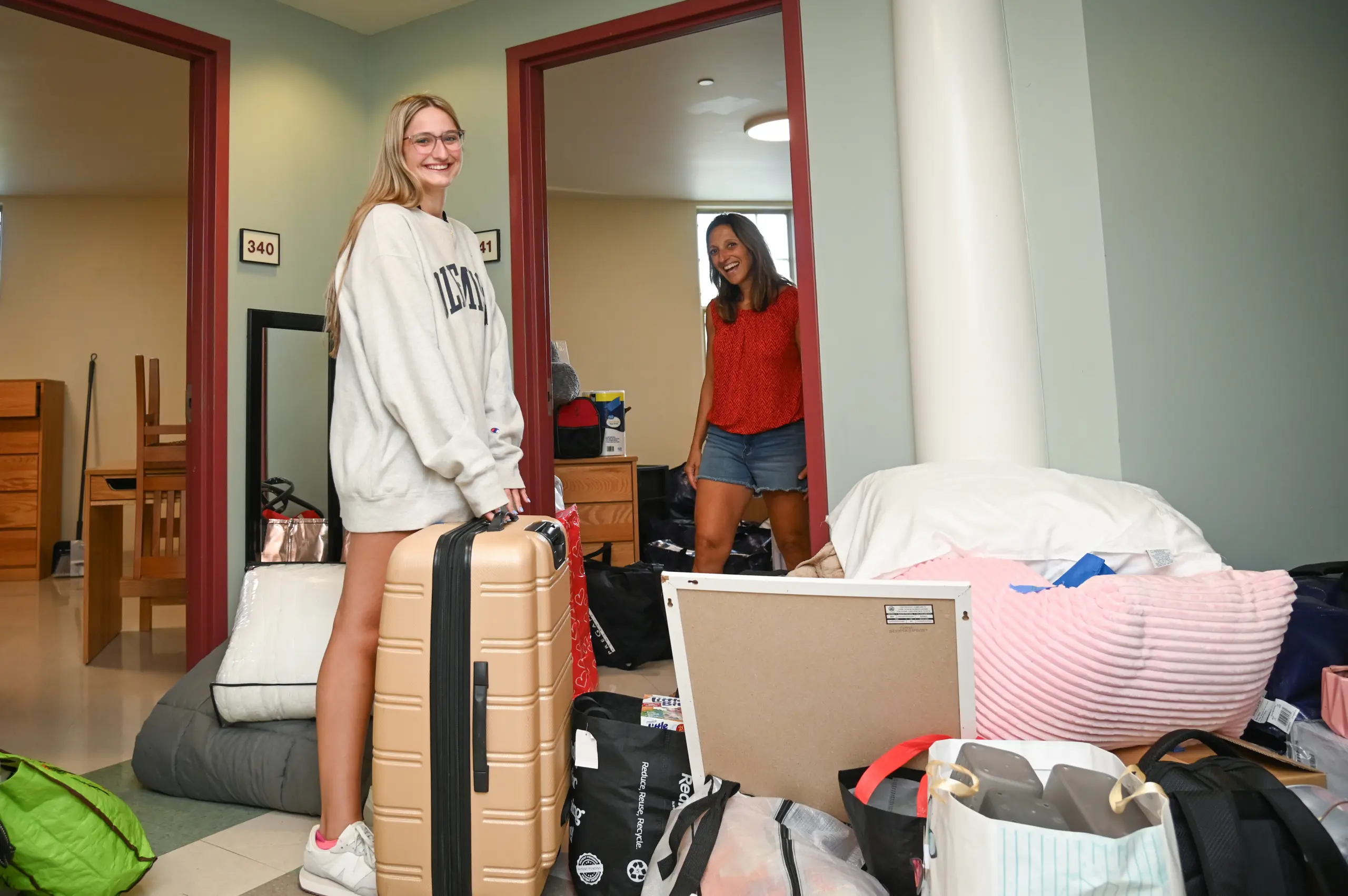 A young woman in a sweatshirt stands in a dorm hallway with a suitcase, smiling at the camera. Another woman stands in a doorway behind her. Bags, bedding, and supplies are piled around them, capturing the excitement of First Year Housing move-in day.