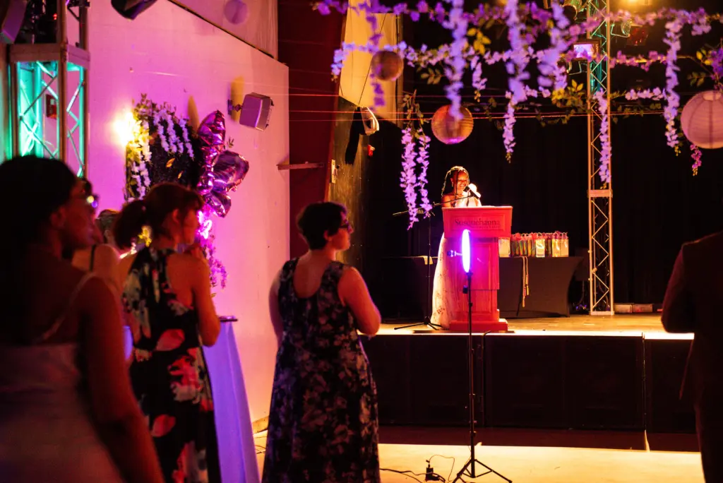 A woman stands at a podium speaking to an audience in a dimly lit event space decorated with flowers, hanging lanterns, and lights. Several people in formal attire listen attentively.