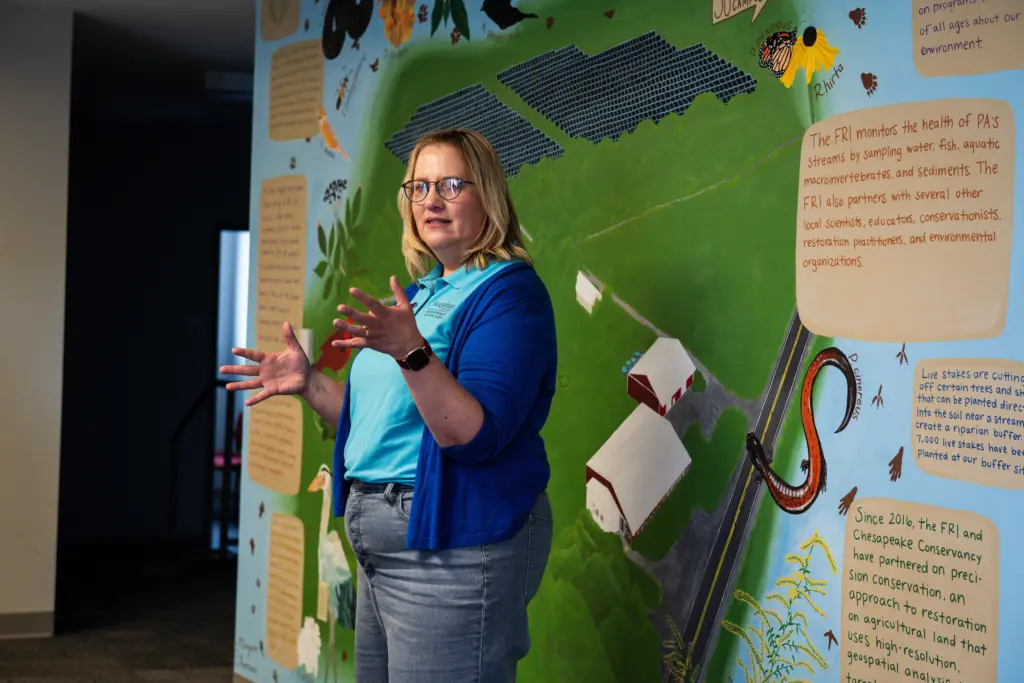 A woman with glasses gestures while speaking in front of a colorful educational mural featuring farms, fields, and text about environmental research, conservation, and research opportunities.