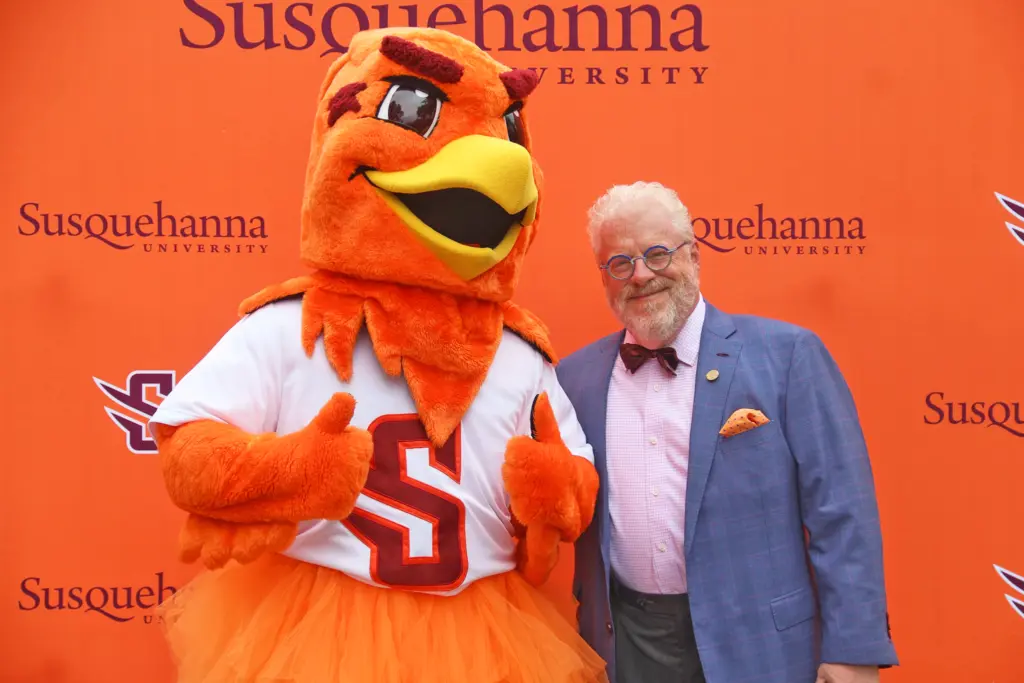 Our Leadership shines as a person in a blue suit and bow tie stands smiling next to an orange bird mascot in front of a 体育买球 University backdrop. The mascot gives a thumbs up, wearing an 