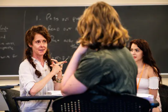 A woman with braided hair speaks to two students in a classroom, gesturing with her hands as she discusses academics. A chalkboard with writing is visible in the background while the students listen attentively.