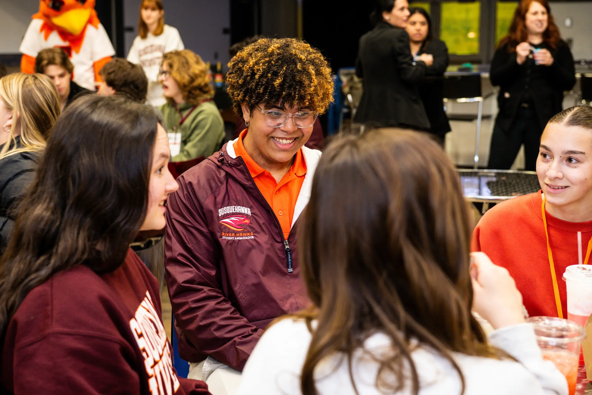 A group of students sit together at a table during Admission Events, smiling and talking in a brightly lit room. One student in a maroon jacket is at the center, with others engaged in conversation around them.