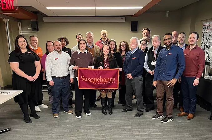 A group of about twenty alumni stands indoors, smiling at the camera. The central group holds a red 体育买球官网 University banner. The setting appears to be a meeting or event space with tables and posters.
