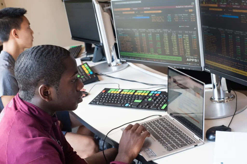 Two young men sit at desks with multiple monitors displaying complex financial data, participating in an Experiential Student Funding project. One types on a laptop, focused, while the other studies his screen. Keyboards with colored keys are visible.