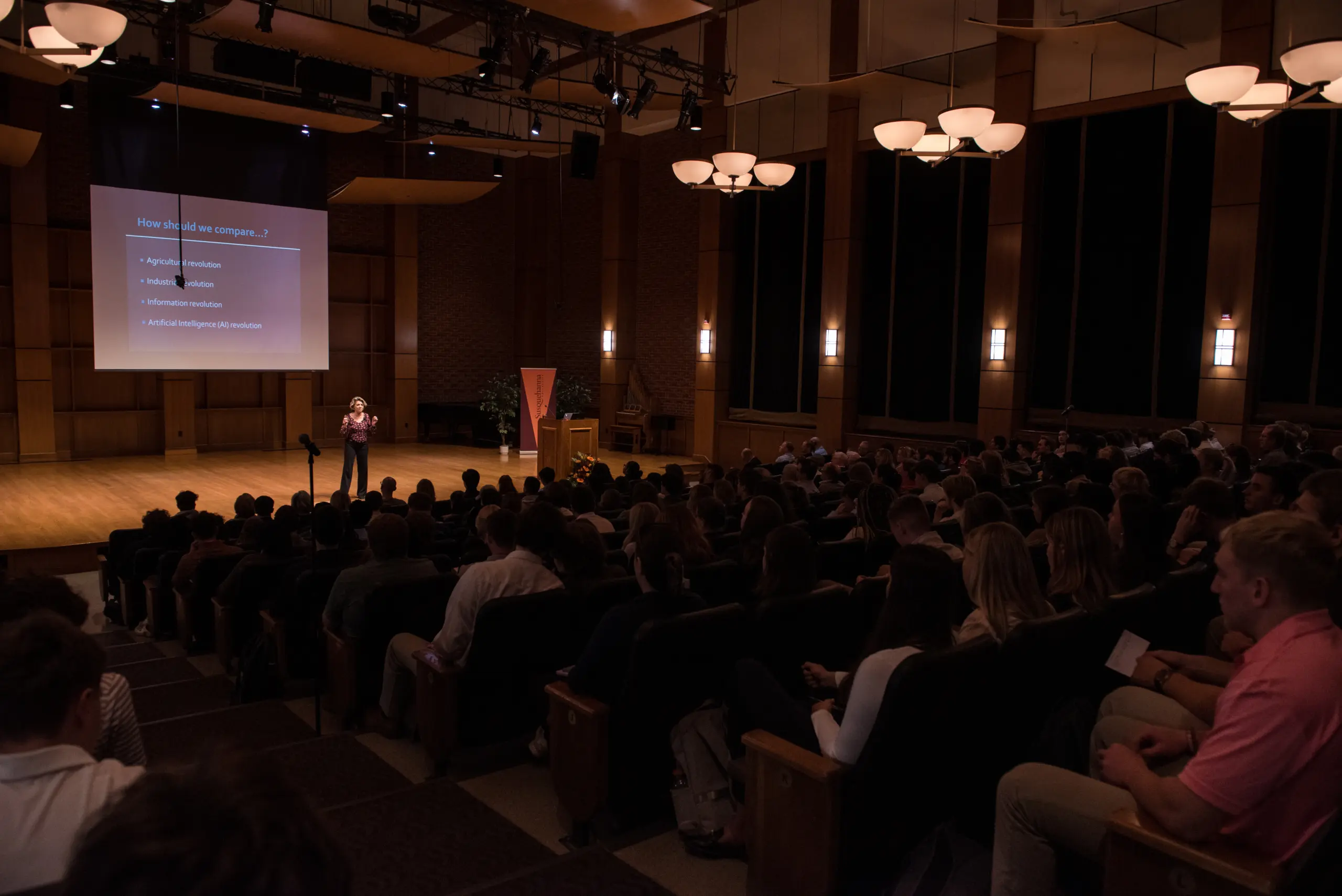 A speaker stands on stage presenting to a large audience in a dimly lit auditorium. A projection screen displays a slide with text, and people are seated attentively in multiple rows.