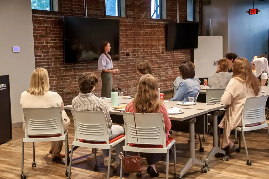 A woman stands and speaks in front of a group seated at tables in a meeting room with exposed brick walls, large monitors, and windows, fostering Community Access as the audience attentively listens.