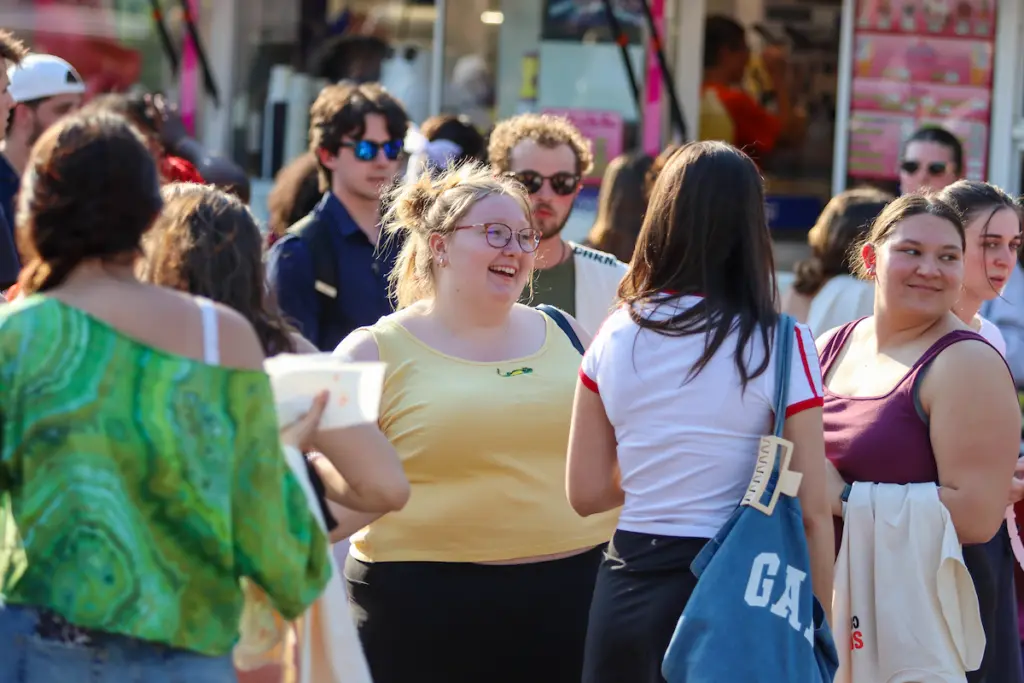A group of people stand and converse outdoors on a sunny day, possibly discussing admission details. One person in a yellow tank top and glasses smiles at others, with a busy storefront and blurred shoppers in the background.