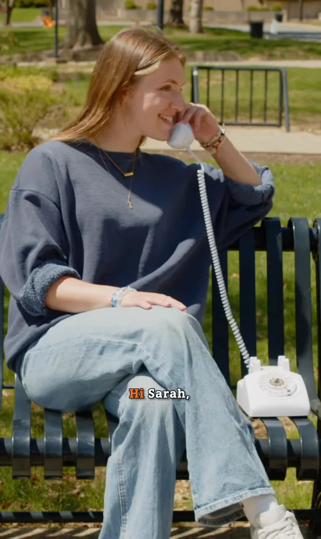 A young woman sits on a park bench, smiling while talking on a white rotary phone. She wears a loose blue sweater and light jeans, celebrating her commencement moment. The subtitle says, 