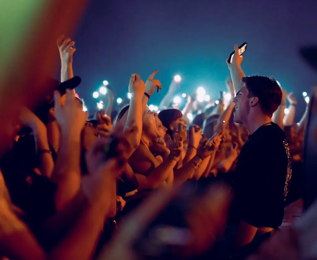 A crowd with admission eagerly raises their hands and phones with flashlights at a concert, as a person in the front row sings or cheers, illuminated by stage lights.