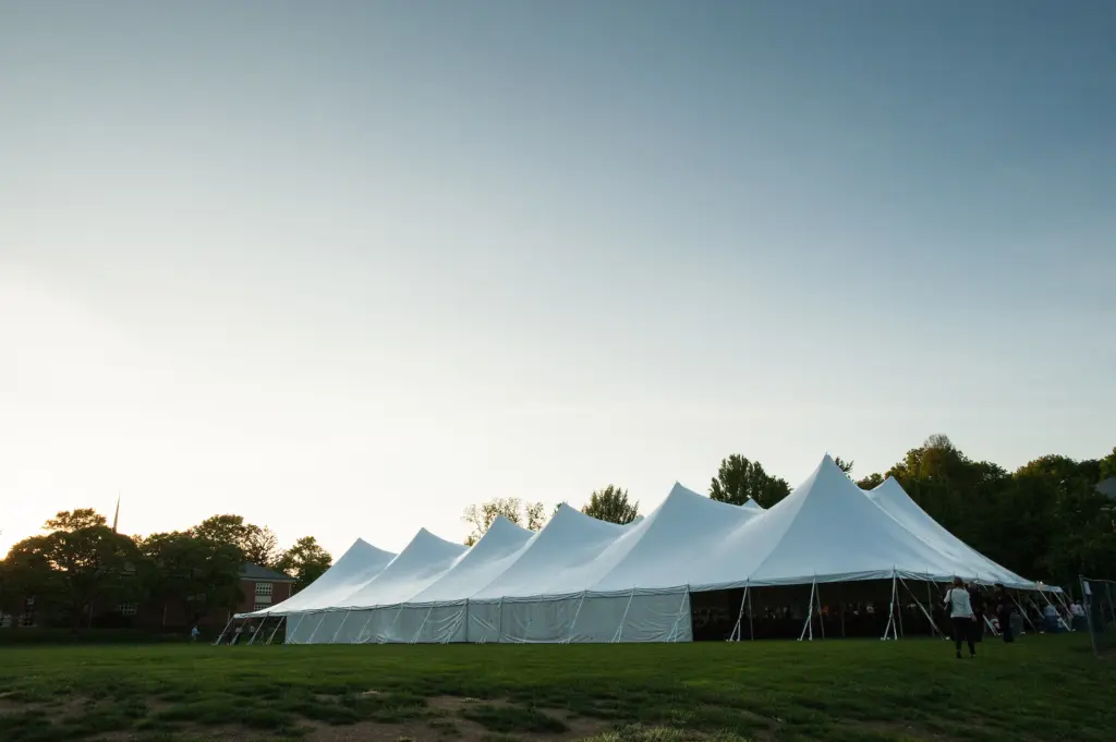 A large white event tent with multiple peaks is set up on a grassy field at dusk, with trees and a few people visible around the tent. The sky is clear and fading to evening.
