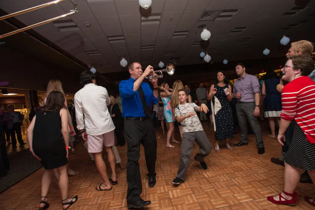A lively indoor party scene with people dancing on a wooden floor; a man in a blue shirt plays a trumpet while a child in patterned clothing dances enthusiastically nearby. Others stand and watch, smiling and talking.