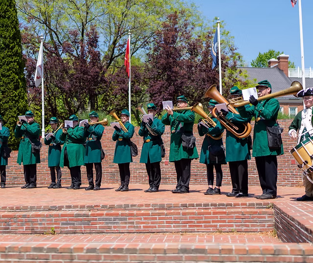 A marching band in green uniforms plays brass instruments outdoors on brick steps, celebrating community access to music. Flags and trees frame the scene, with a drummer in a white uniform standing to the right.