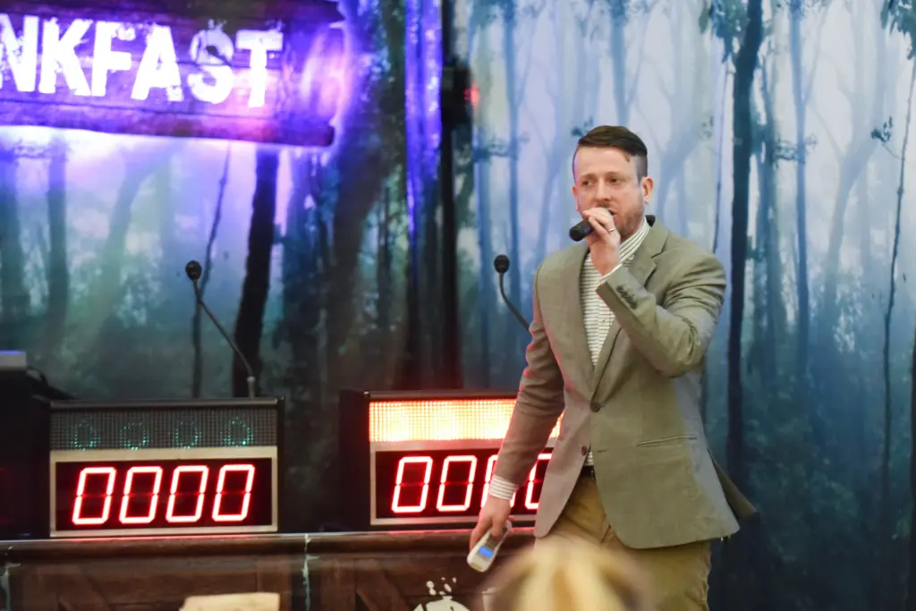 A man in a beige blazer holds a microphone and speaks on stage in front of two scoreboard displays showing zeros, with a forest-themed backdrop and a partially visible illuminated sign reading “NKFAST.”.