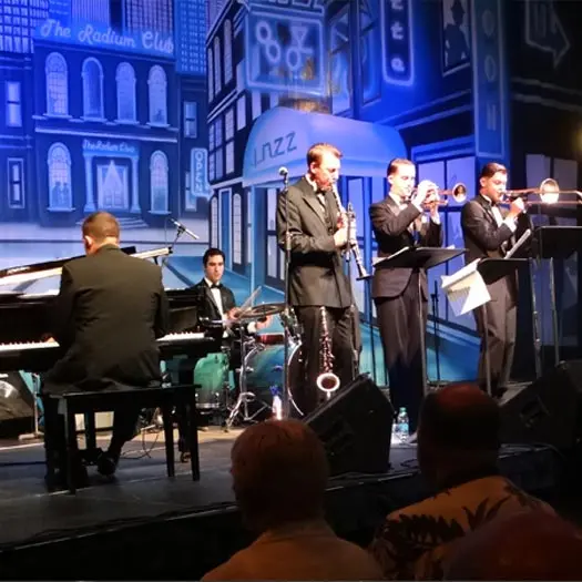 A jazz band in suits performs on stage with piano, drums, clarinet, trumpet, and saxophone; the backdrop features blue-toned city buildings and “jazz” signs, celebrating community access as an audience watches.