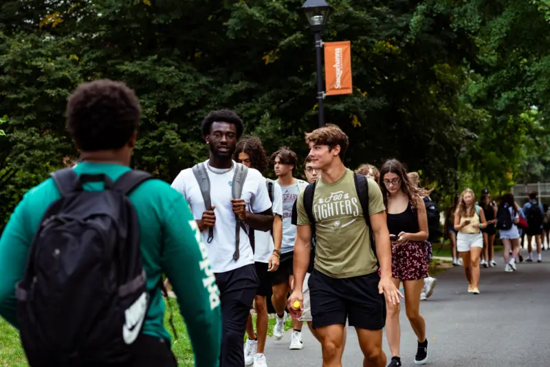 A group of college students walk along a tree-lined campus path. Some carry backpacks, and a few are talking or looking at their phones. A university banner hangs on a lamp post in the background.