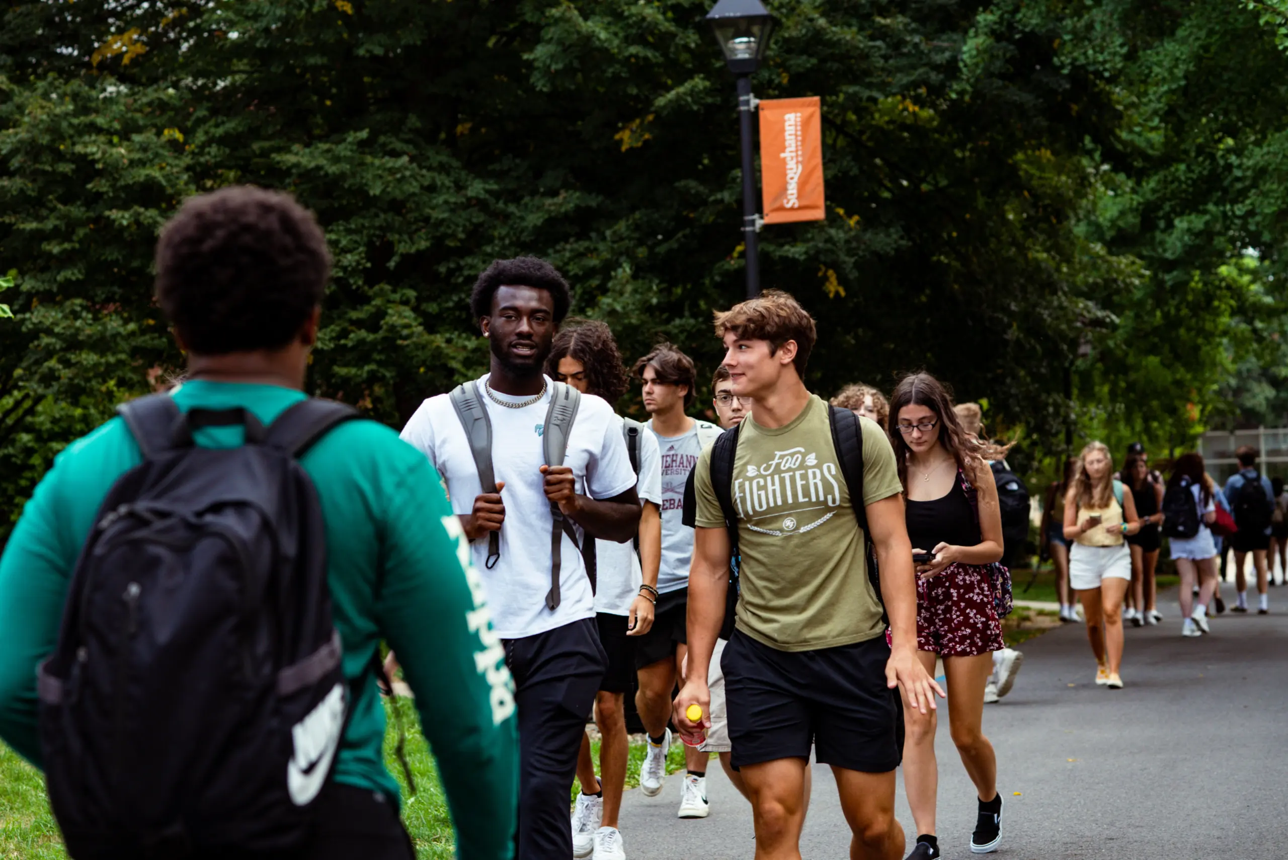 A group of college students walk along a tree-lined campus path. Some carry backpacks, and a few are talking or looking at their phones. A university banner hangs on a lamp post in the background.