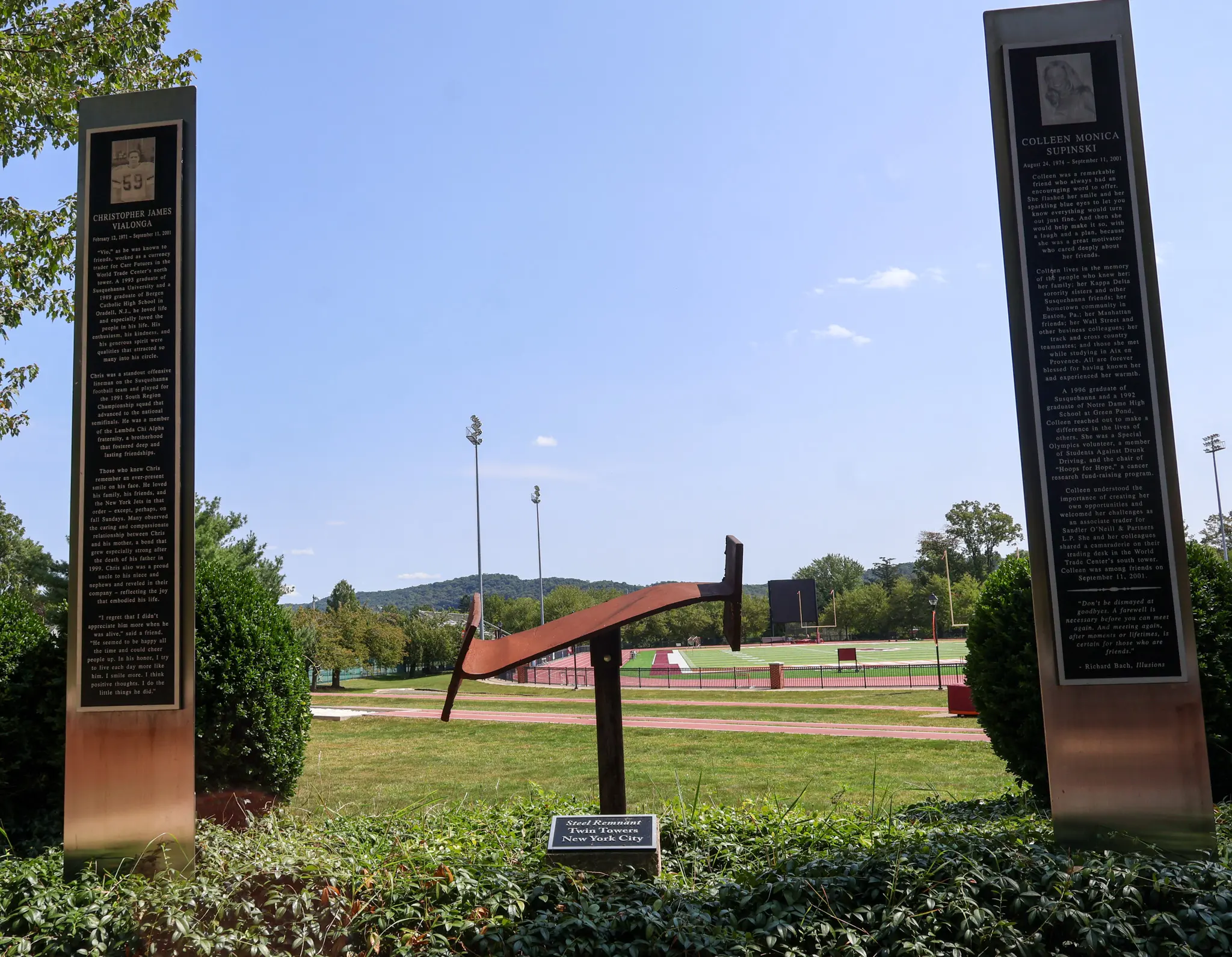 Two tall plaques with text and images stand on either side of a metal sculpture shaped like a plow, set on a grassy area with a sports field and trees visible in the background under a clear sky.