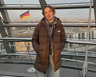 A person wearing a brown winter coat and scarf stands indoors with their hands in their pockets. Behind them, a German flag is visible outside against a cityscape.