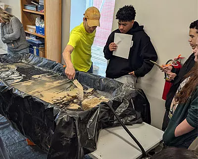 A man in a yellow shirt demonstrates a sand and rock model to a group of students, some holding notebooks and one holding a fire extinguisher, in a classroom setting.