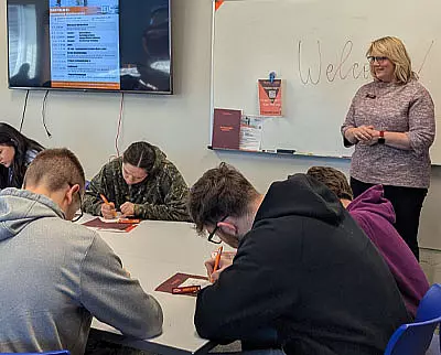 A woman stands and speaks in front of a whiteboard that says “Welcome,” while four students sit at a table, writing in notebooks. A screen with a document is displayed on the wall in the background.