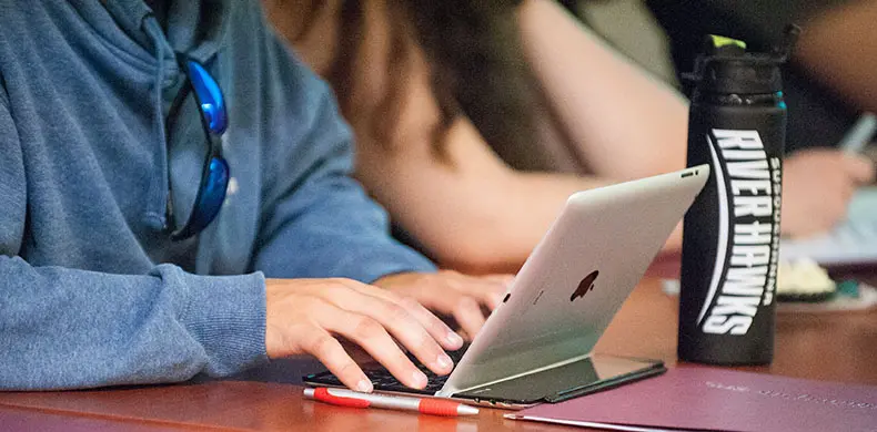 A person wearing a blue hoodie types on a silver laptop at a desk. A black water bottle labeled 
