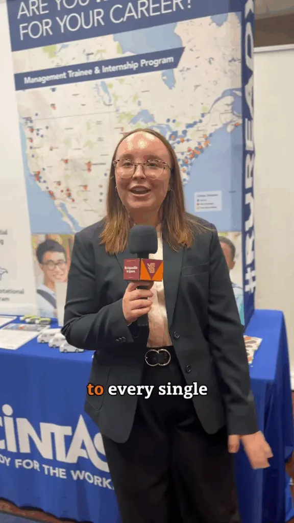 A person in a black blazer is standing with a microphone in front of a booth with a banner that says 