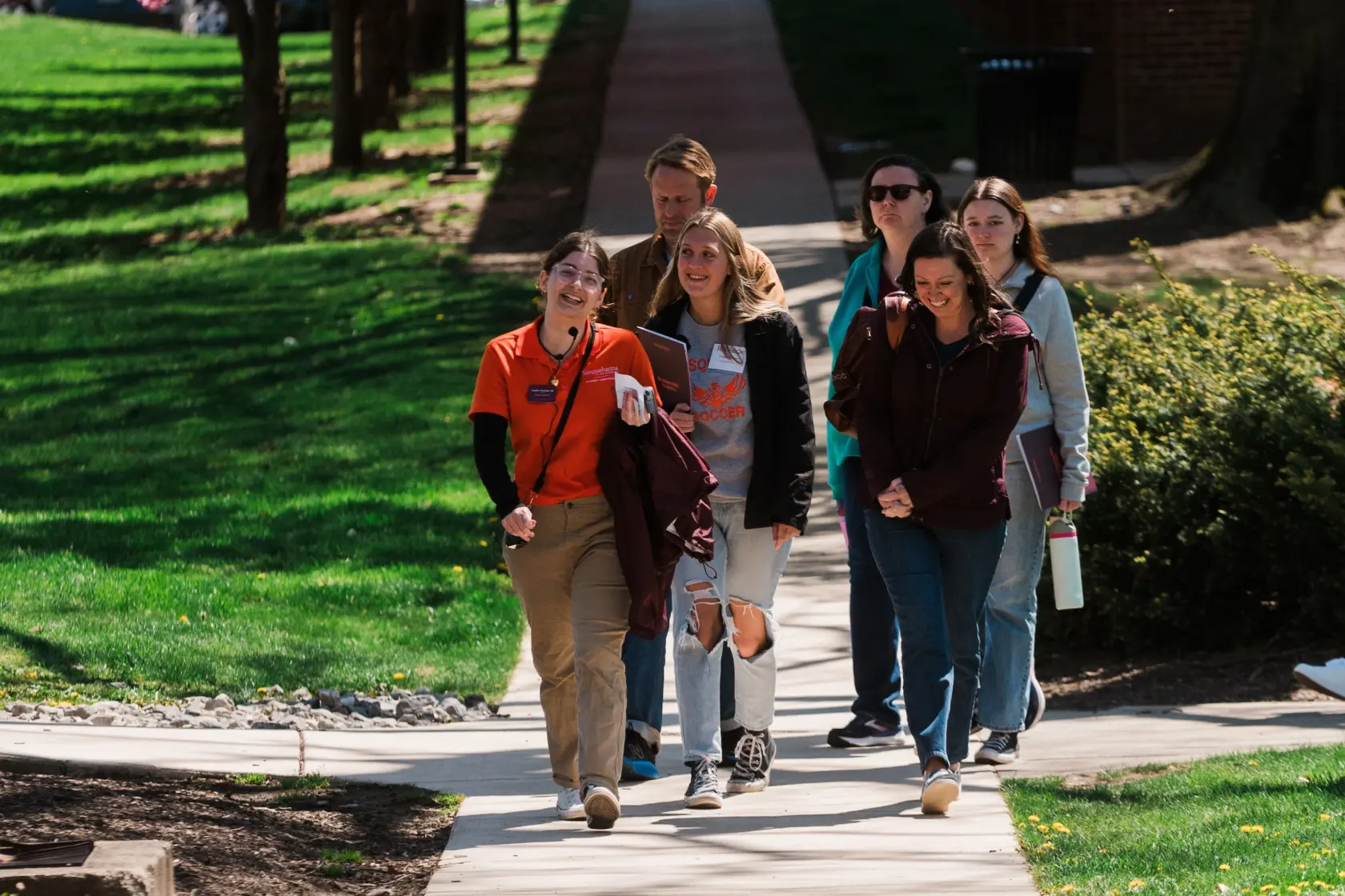 A group of seven people, some wearing university name tags and carrying water bottles, walk together on a sunny campus sidewalk surrounded by grass and trees.