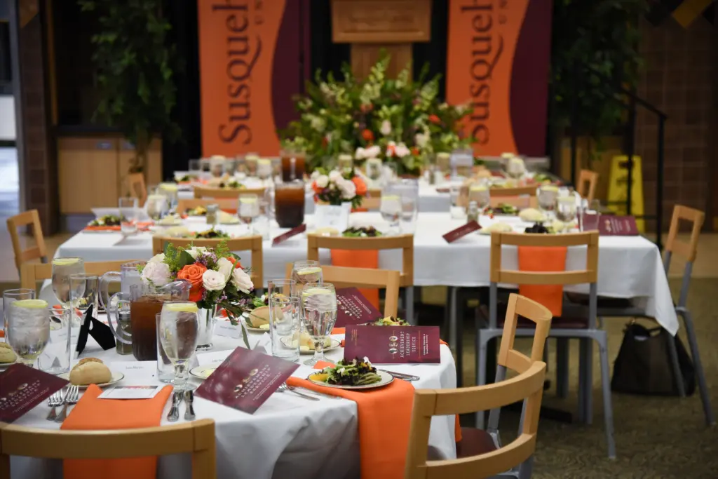 A banquet setup with tables covered in white cloths, orange napkins, flower centerpieces, salads, drinks, and menus. In the background, banners reading “体育买球官网,” a large floral arrangement, and signs for Conference Facility Rental are visible.