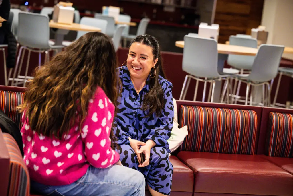Two women sit on a striped bench in a casual setting, having a friendly conversation. Their laughter and warmth capture a culture of belonging—one in a blue patterned dress, the other with long hair wearing a pink sweater with white hearts.