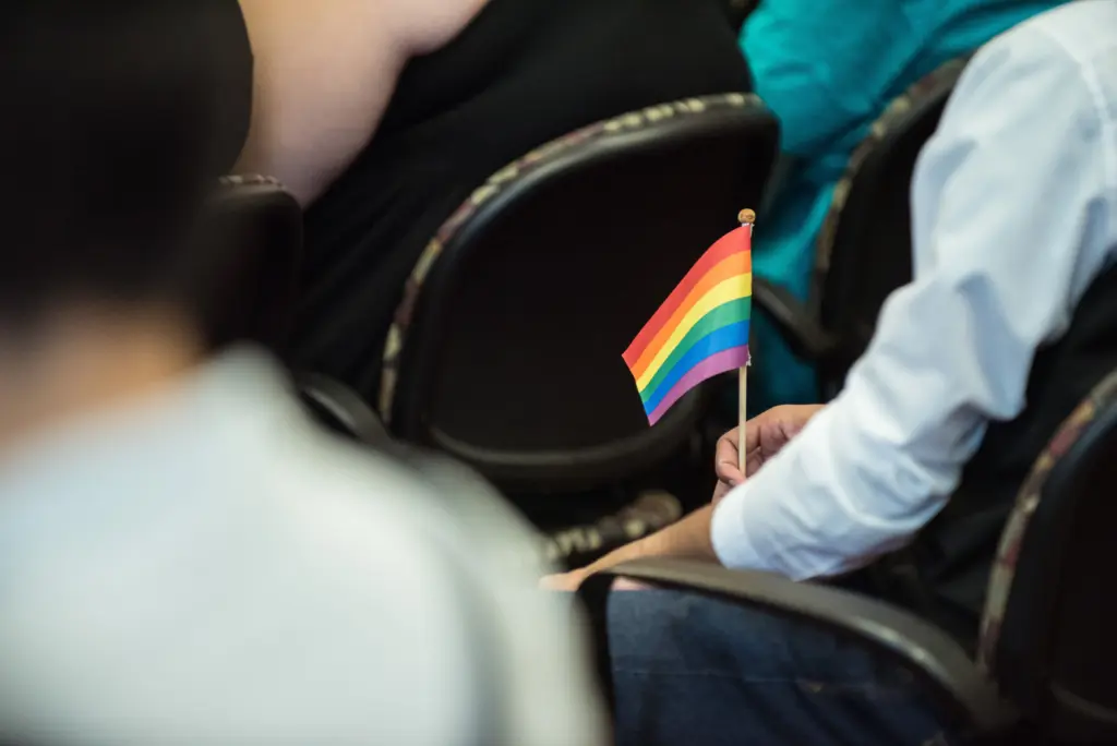 A person seated among others holds a small rainbow pride flag, symbolizing the LGBTQ+ community's pride, in a room lined with rows of chairs.