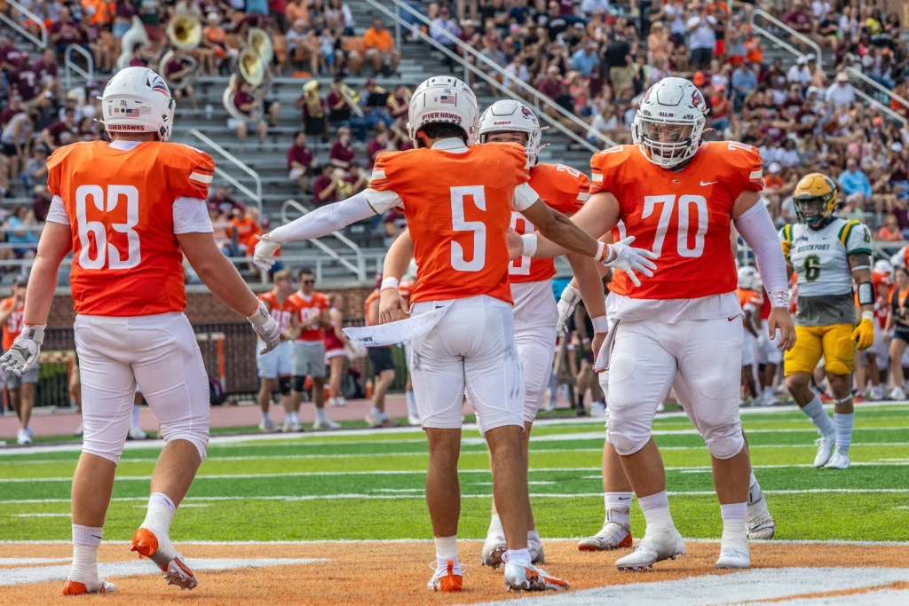 Four football players in orange jerseys celebrate on the field during a game, with one holding out his arms. The excitement of Homecoming-Reunion fills the air as fans and opposing team members look on in the background.